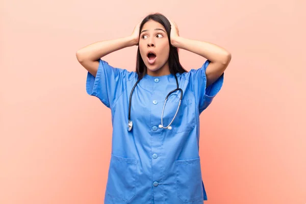 young latin nurse with open mouth, looking horrified and shocked because of a terrible mistake, raising hands to head against pink wall