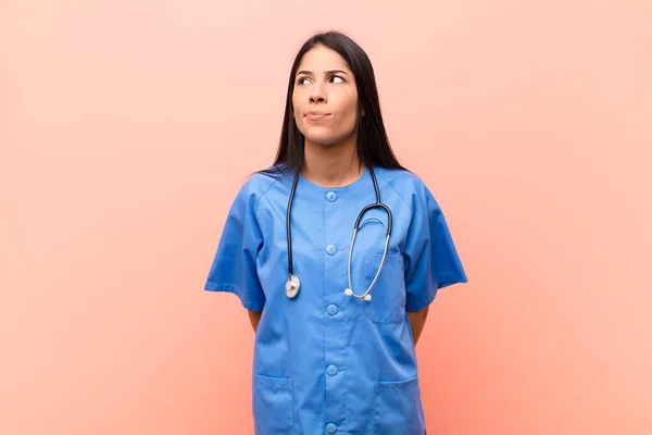 young latin nurse looking puzzled and confused, wondering or trying to solve a problem or thinking against pink wall