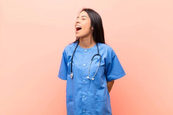 young latin nurse screaming furiously, shouting aggressively, looking stressed and angry against pink wall