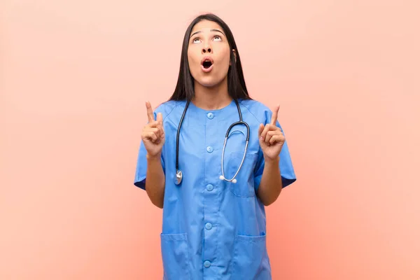 young latin nurse feeling awed and open mouthed pointing upwards with a shocked and surprised look against pink wall