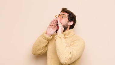 Havalı genç adam mutlu, heyecanlı ve pozitif hissediyor. Elleri ağızda, düz duvara karşı bağırıyor.