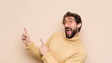 Genç havalı adam mutlu bir şekilde gülümsüyor ve iki eliyle yukarı ve yana doğru işaret ediyor fotokopi alanında düz duvara karşı nesne gösteriyor.