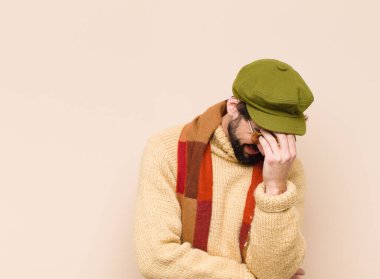 Genç havalı sakallı adam stresli, utanmış ya da üzgün görünüyor, baş ağrısı var, yüzünü eliyle kapatıyor.