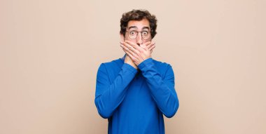 Genç yakışıklı adam elleri ile ağzını kapatıyor, şok olmuş, şaşırmış bir ifadeyle, bir sır saklıyor ya da düz duvara karşı 