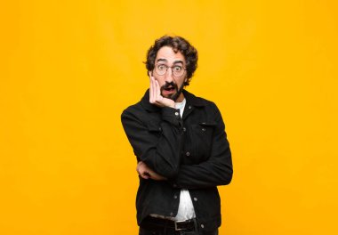 Genç çılgın yakışıklı adam şok ve inançsızlık içinde, elleri yanak ve kolları çaprazlanmış, sersemlemiş ve turuncu duvara karşı şaşkın hissediyordu.