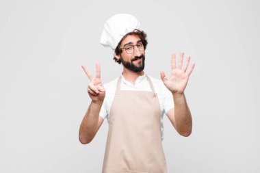 Genç ve çılgın şef gülümsüyor ve dost canlısı görünüyor. Elleri önde, beyaz duvara doğru geri sayarak yedi ya da yedinci numarayı gösteriyor.