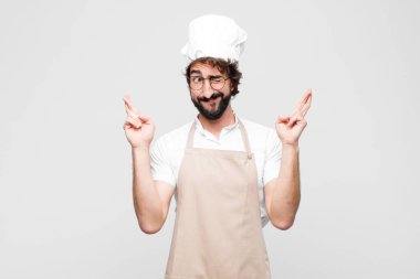 Genç deli şef gülümsüyor ve endişeyle iki parmağını çapraz yapıyor. Endişeli hissediyor ve beyaz duvara karşı iyi şanslar diliyor veya umut ediyor.