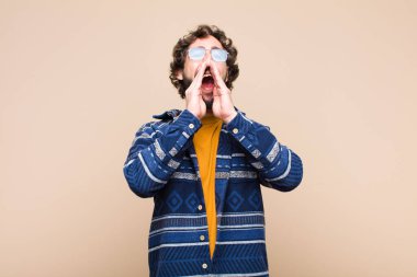 Genç, çılgın, havalı adam mutlu, heyecanlı ve pozitif hissediyor. Elleri ağızda, düz duvara karşı bağırıyor.