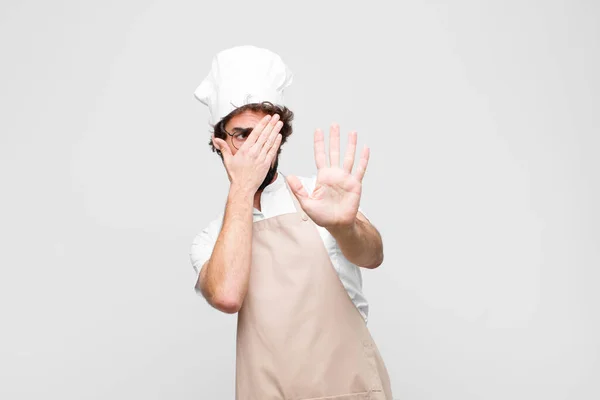 young crazy chef covering face with hand and putting other hand up front to stop camera, refusing photos or pictures against white wall