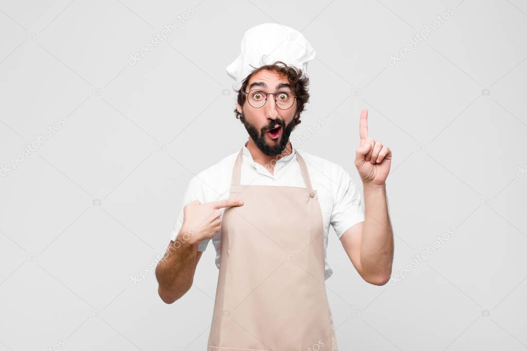 young crazy chef feeling proud and surprised, pointing to self confidently, feeling like successful number one against white wall