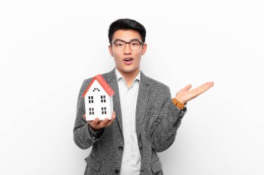 Genç japon adam şaşırmış ve şok olmuş görünüyor, çenesi düşmüş, eli açık bir objeyi elinde tutarken bir ev modeliyle