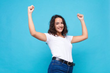 Genç ve güzel bir kadın muzaffer bir şekilde bağırıyor, heyecanlı, mutlu ve şaşırmış görünüyor, mavi arka planda kutlama yapıyor.