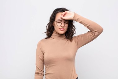 Genç ve güzel bir kadın stresli görünüyor, yorgun ve hayal kırıklığına uğramış, alnından ter damlıyor, umutsuz ve yorgun hissediyor beyaz duvara karşı.