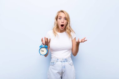 Genç sarışın kadın şok olmuş, şaşırmış, endişeli ve paniklemiş hissediyor. Stresli ve dehşetli bir bakışla elinde bir saat tutuyor.