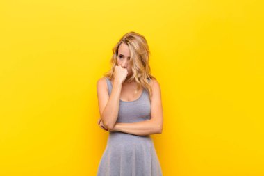 Genç sarışın kadın ciddi, düşünceli ve endişeli. Eli turuncu duvara yaslanmış bir şekilde yan bakıyor.