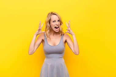 Genç sarışın kadın ellerini havaya kaldırarak bağırıyor. Öfkeli, sinirli, gergin ve turuncu duvara karşı üzgün.