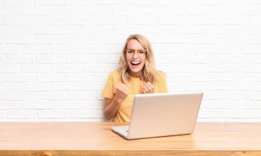 Genç sarışın kadın sinirli sinirli, sinirli bakışlar ve yumruklarla bağırıyor. Dizüstü bilgisayar kullanarak öfkeleniyor.