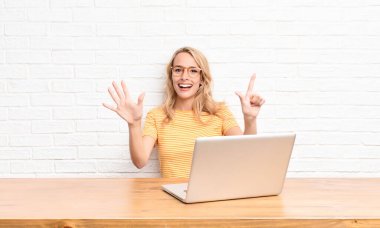 Genç sarışın kadın gülümsüyor ve arkadaş canlısı görünüyor. 7 ya da 7 numarayı gösteriyor. Elleri önde, dizüstü bilgisayarla geri sayıyor.