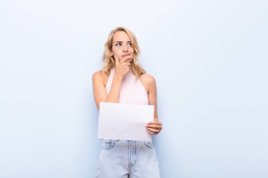 Genç sarışın kadın düşünüyor, şüpheci ve şaşkın hissediyor, farklı seçenekleri var, elinde bir kağıt tutarak hangi kararı vereceğini merak ediyor.