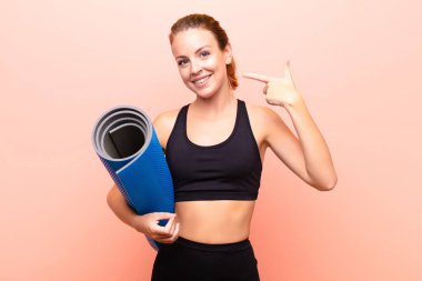 Kızıl saçlı, güzel, kendine güvenen, gülümseyen, pozitif, rahat, tatmin olmuş, yoga minderi tutan bir kadın..