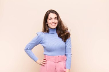 Genç ve güzel bir kadın mutlu bir şekilde gülümsüyor, eli kalçasında ve kendinden emin, pozitif, gururlu ve arkadaş canlısı bir tavırla bej arka plana karşı.