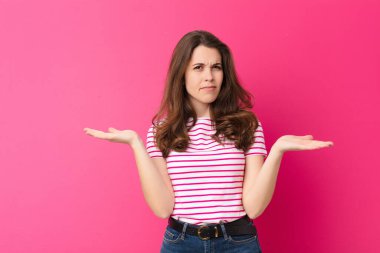 Genç, güzel bir kadın şaşkın, kafası karışmış ve stresli görünüyor, farklı seçenekler arasında merak ediyor, pembe duvara karşı kararsız hissediyor.