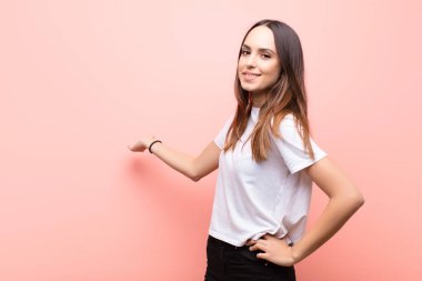 Genç, güzel bir kadın kendini mutlu ve neşeli hissediyor, gülümsüyor ve seni hoş karşılıyor, pembe duvara karşı dostça bir jestle içeri davet ediyor.