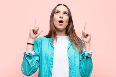 Genç, güzel bir kadın şaşırmış, şaşkın ve ağzı açık görünüyor, iki eliyle yukarıya bakıyor pembe duvara uzayı kopyalamak için.