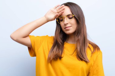 Genç ve güzel bir kadın stresli görünüyor, yorgun ve hayal kırıklığına uğramış, alnından ter damlıyor, mavi duvara karşı umutsuz ve bitkin hissediyor.