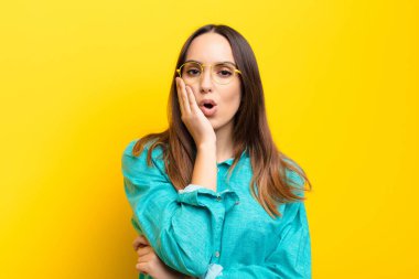 Genç, güzel bir kadın şok olmuş ve şok olmuş hissediyor. Ağzı açık bir şekilde turuncu duvara dayanmış, yüz yüze inanamıyor.