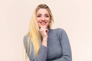 Genç ve güzel sarışın kadın mutlu görünüyor ve gülümsüyor, çenesi açık, merak ediyor ya da soru soruyor, düz duvarla seçenekleri karşılaştırıyor.