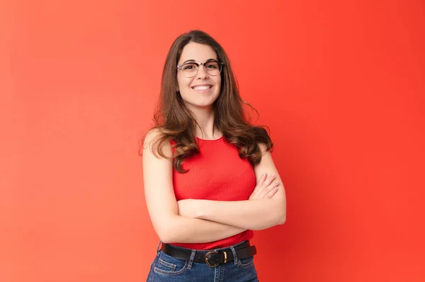 young pretty woman smiling to camera with crossed arms and a happy, confident, satisfied expression, lateral view against red wall