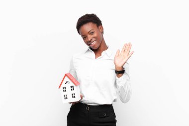 Genç ve güzel siyah kadın mutlu ve neşeyle gülümsüyor, el sallıyor, sizi selamlıyor ve selamlıyor, ya da bir ev modeliyle vedalaşıyor.