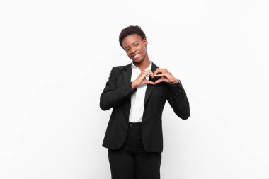 Genç ve güzel siyah kadın gülümsüyor ve mutlu, sevimli, romantik ve aşık, iki elini de beyaz duvara dayayarak kalbi şekillendiriyor