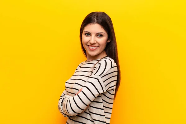 young pretty woman smiling to camera with crossed arms and a happy, confident, satisfied expression, lateral view against yellow wall