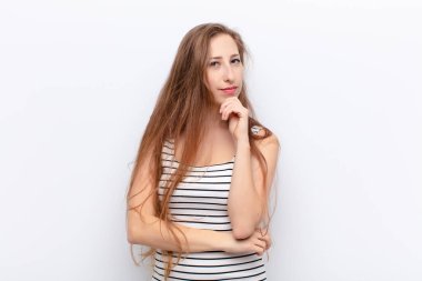 Genç sarışın kadın ciddi görünüyor, kafası karışık, kararsız ve düşünceli, beyaz duvara karşı seçenekler veya seçimler arasında şüpheci