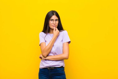 Genç, güzel, Latin bir kadın ciddi görünüyor, kafası karışık, kararsız ve düşünceli, seçenekler arasında veya düz duvara karşı seçimler yapıyor.