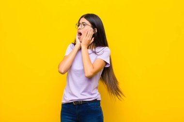 Genç, güzel, Latin bir kadın mutlu, heyecanlı ve şaşırmış hissediyor. İki eli yüzü düz duvara dönük bir şekilde yan tarafa bakıyor.