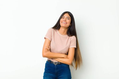 Genç, güzel, Latin bir kadın kollarını kavuşturup mutlu bir şekilde gülüyor, rahat, pozitif ve tatmin olmuş bir pozla düz duvara yaslanıyor.