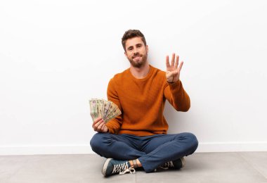 Genç yakışıklı adam gülümsüyor ve arkadaş canlısı görünüyor, dört ya da dört numarayı gösteriyor, elinde dolar banknotları tutarak geriye doğru sayıyor.