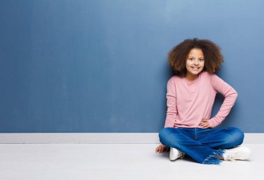 Afro-Amerikan küçük kızı mutlu bir şekilde gülümsüyor elleri kalçasında ve kendinden emin, olumlu, gururlu ve arkadaş canlısı bir tavırla yerde oturuyor.