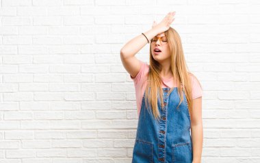 young blonde woman raising palm to forehead thinking oops, after making a stupid mistake or remembering, feeling dumb against brick wall