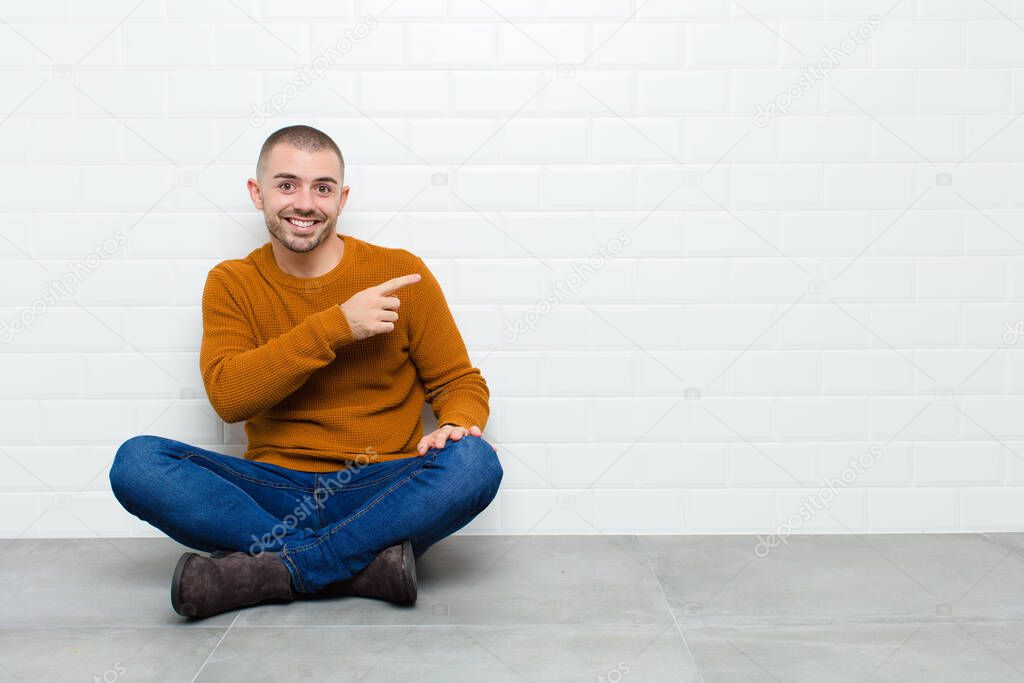 young handsome man looking excited and surprised pointing to the side and upwards to copy space sitting on the floor