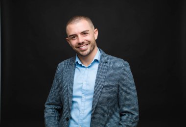 Genç işadamı neşeli bir şekilde gülümsüyor. Olumlu, mutlu, kendinden emin ve gevşek bir ifadeyle düz duvara yaslanıyor.