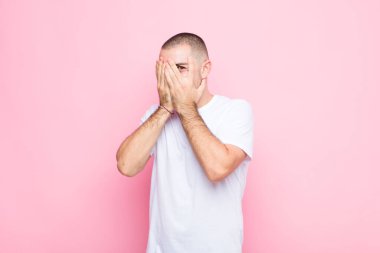 Genç yakışıklı adam elleriyle yüzünü kapatıyor, parmaklarının arasından şaşkın bir ifadeyle bakıyor ve düz duvara bakıyor.