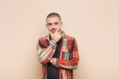 Genç yakışıklı adam ciddi, düşünceli ve güvensiz görünüyor, bir kolu çapraz ve çene üzerinde, düz duvara karşı seçenekleri tartıyor