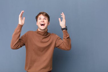 Genç sarışın adam kendini mutlu, şaşkın, şanslı ve şaşırmış hissediyor, zaferi kutluyor, iki eli havada, düz duvara karşı izole bir şekilde.