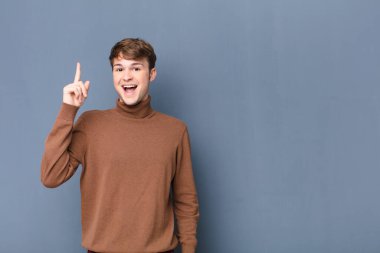 Genç sarışın bir adam bir fikri fark ettikten sonra mutlu ve heyecanlı bir dahi gibi hissediyor, neşeli bir şekilde parmağını kaldırıyor, eureka! Düz duvara karşı izole