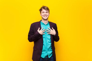 Genç sarışın adam mutlu, şaşırmış, gururlu ve heyecanlı görünüyor. Kendini düz duvara yaslamış gösteriyor.