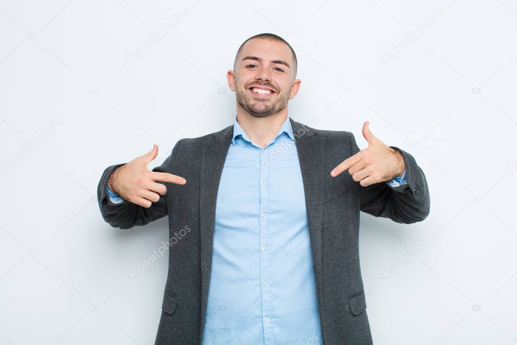 young businessman looking proud, arrogant, happy, surprised and satisfied, pointing to self, feeling like a winner against flat wall